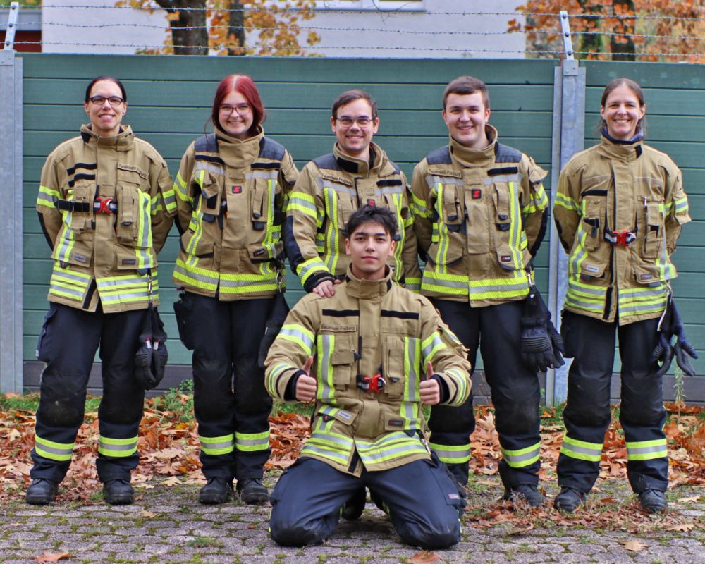 Gruppenfoto der sechs Teilnehmer am Grundlehrgang, sie stehen in Feuerwehrkleidung in einer Reihe und lachen in die Kamera.