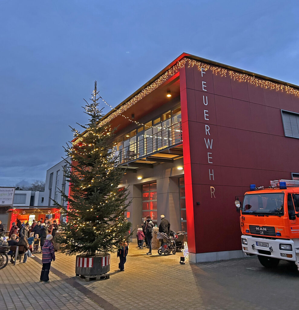 Ansicht aufs beleuchtete Gerätehaus mit Weihnachtslichtern, rechts ist ein Feuerwehrfahrzeug zu sehen, in der MItte ein Weihnachtsbaum und einige Besucher.