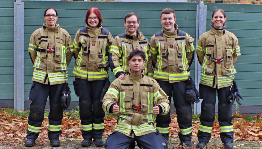 Gruppenfoto der sechs Teilnehmer am Grundlehrgang, sie stehen in Feuerwehrkleidung in einer Reihe und lachen in die Kamera.